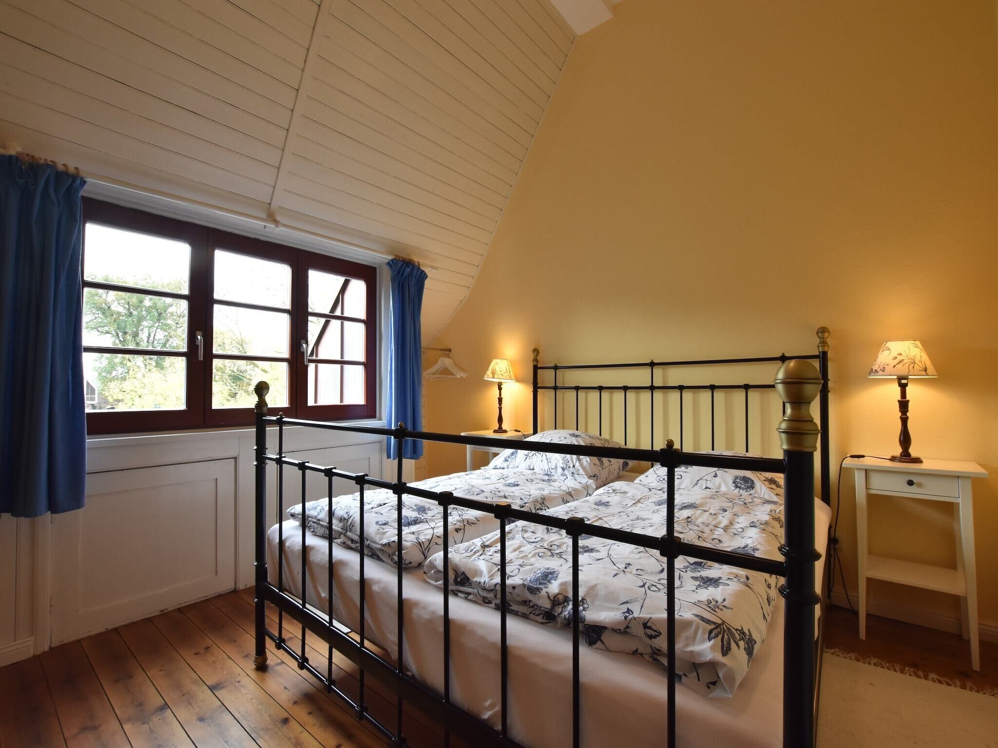 Un dormitorio con techo inclinado, una cama con estructura de metal, dos mesitas de noche con lámparas y una ventana con cortinas.