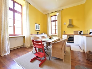 A bright yellow room with a dining table, wicker chairs, a red chair, a kitchen with white cabinets, a stove, and a clock on the wall. - Wohnung in Satow mit Garten (Gerdshagen)