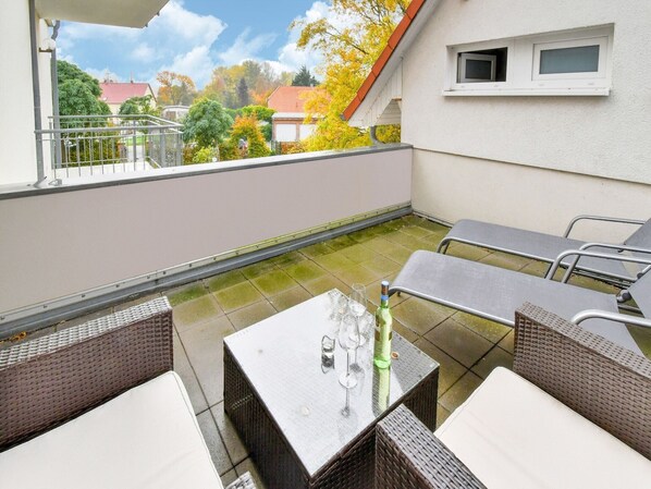 An outdoor patio with wicker furniture, a table with a bottle and glasses, and a view of houses and trees.