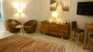 Habitación de hotel con una cama, dos sillones, un aparador de madera, un escritorio con una silla, un televisor de pantalla plana y luces fijadas a la pared.