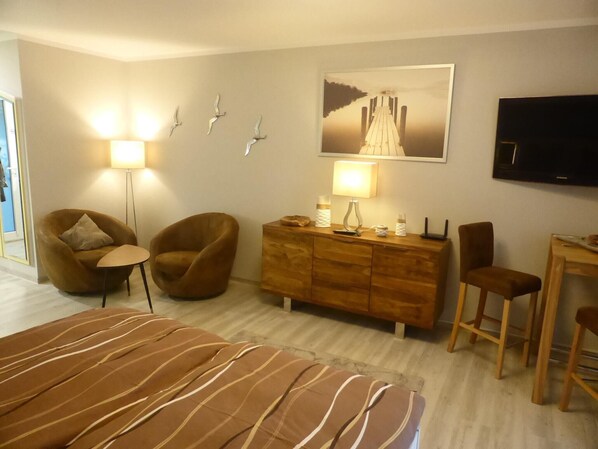 A hotel room with a bed, two armchairs, a wooden dresser, a desk with a chair, a flat-screen TV, and wall-mounted lights. - Modernes Appartement mit Terrasse auf Rügen (Wiek auf Rügen)