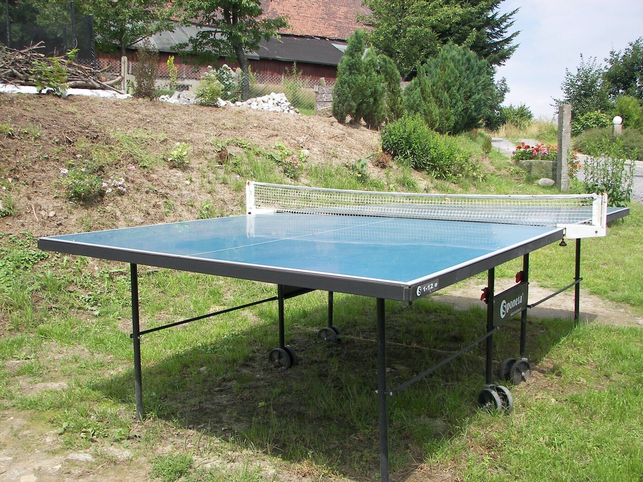 Een buitenpingpongtafel met wielen, opgesteld in een grasveld met bomen en een gebouw op de achtergrond.