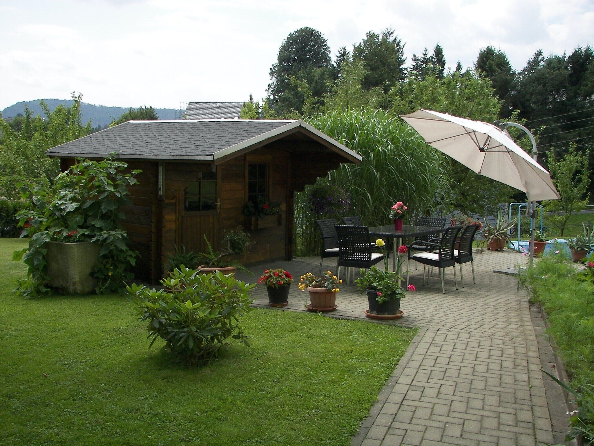 A wooden cabin with a tiled roof, surrounded by a garden with potted plants and a paved patio area with outdoor furniture.