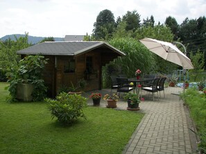 A wooden cabin with a tiled roof, surrounded by a garden with potted plants and a paved patio area with outdoor furniture. - Wohnung in Lichtenhain beim Böhmischen Park (Lichtenhain)