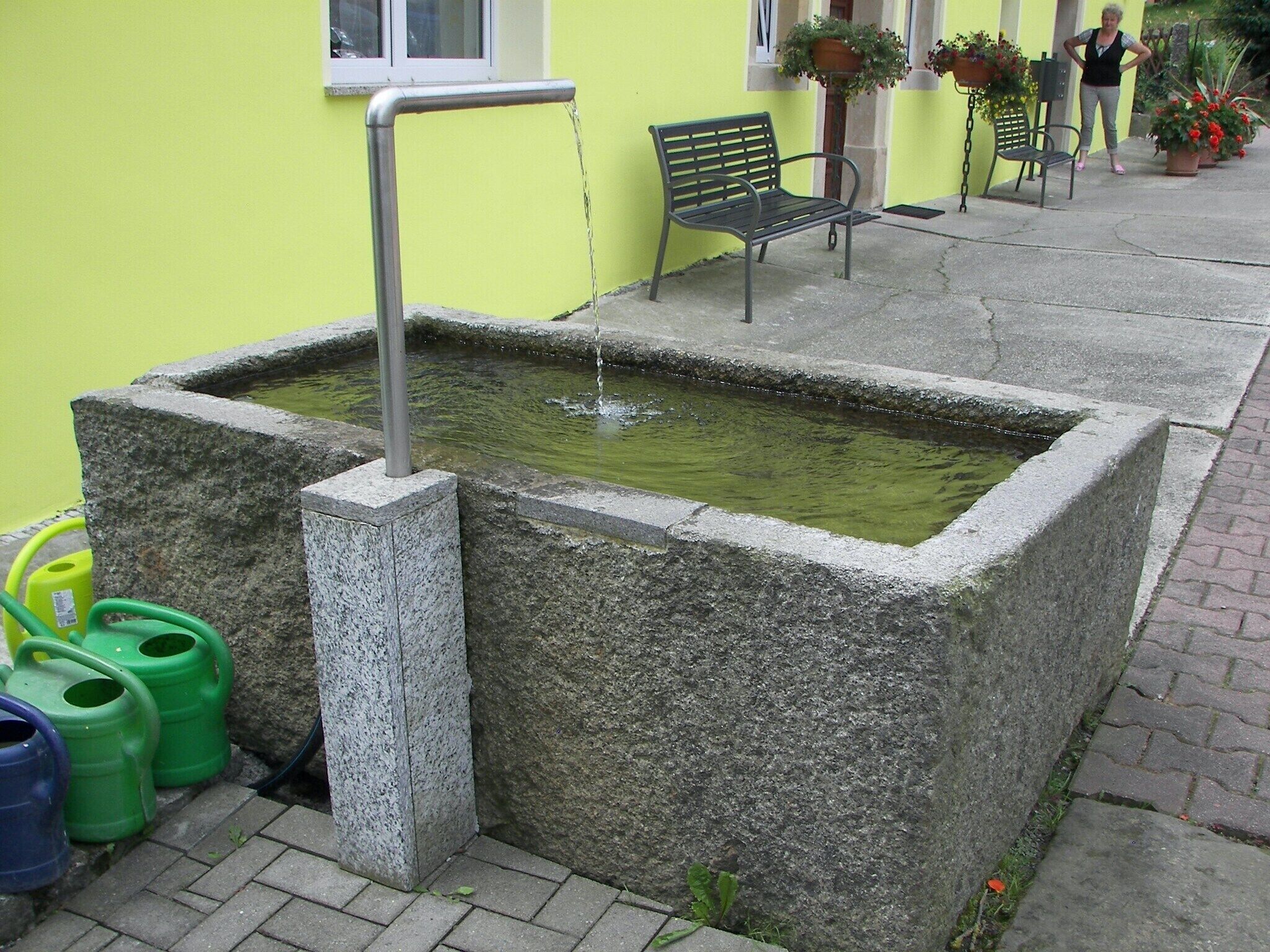 Una fuente pública de agua con un caño metálico, rodeada de bloques de piedra, junto a un banco y plantas en macetas.