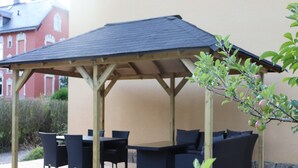 En udendørs terrasse med et trætag, sort udendørs møbler og et lille træ med grønne blade.