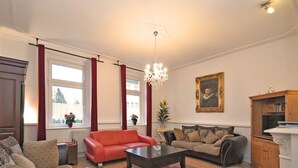 En stue med en rød sofa, en beige sofa, et mørkt sofabord og en lysekrone. Der er vinduer med røde gardiner, et billede på væggen og et træskab.