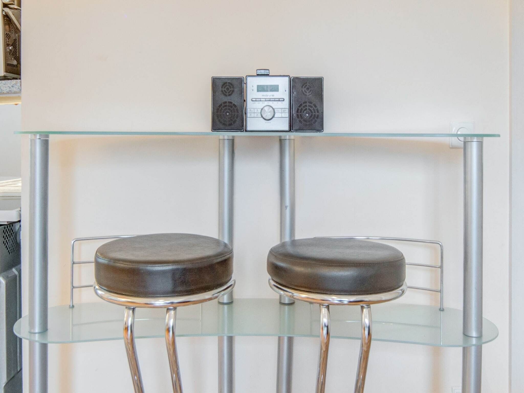 A bar area with two stools and a glass-top table, featuring a digital device and two speakers on the wall.