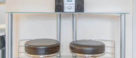 A bar area with two stools and a glass-top table, featuring a digital device and two speakers on the wall.