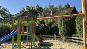 Une aire de jeux avec un toboggan, des balançoires et un petit abri.