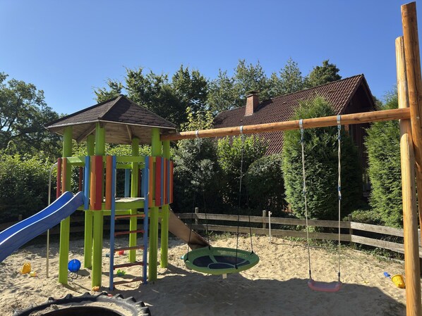 Une aire de jeux avec un toboggan, des balançoires et un petit abri.