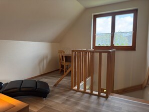 Une pièce avec un plafond en pente, une fenêtre donnant sur un paysage verdoyant, une chaise en bois et un canapé noir.