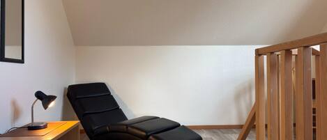 A corner room with a black reclining chair, a desk with a lamp, and a wooden railing.