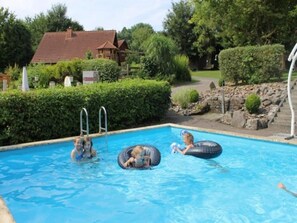 A swimming pool with three people in inflatable rings, surrounded by a landscaped garden and a house in the background. - Holiday Home in Germany with Garden Terrace (Brakel-Bellersen)