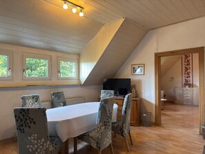 A dining area with a table and chairs, a television, and a view of the outdoors through windows.