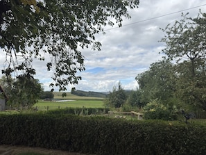 Un paysage rural avec une haie au premier plan, un banc en bois et, au loin, une vue sur un champ et des éoliennes.