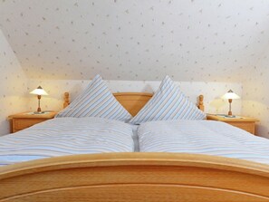 A neatly made bed with striped bedding and two pillows, flanked by wooden nightstands with lamps, in a room with patterned wallpaper and a sloped ceiling. - Wohnung mit Garten in Eimelrod (Willingen-Eimelrod)