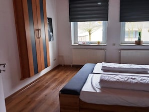 Une chambre avec un plancher en bois, un lit avec une literie blanche, un banc, un radiateur et une fenĂȘtre donnant sur lâextĂ©rieur.