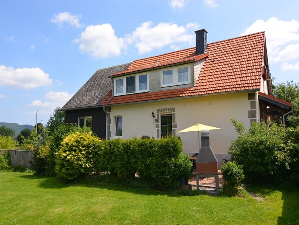 Een woonhuis met twee verdiepingen, voorzien van een rode dakbedekking, een schoorsteen en een klein terras met een barbecue.