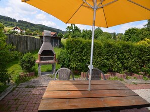 Een terras met een houten tafel, een gele parasol, een stenen barbecue en een rieten stoel.