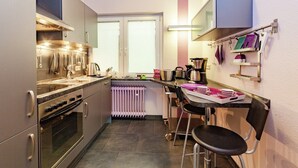 A compact kitchen with a built-in oven, a kettle, and a coffee maker. There is a window with blinds, a radiator, and a small table with a chair. - hundefreundliches Gruppenhaus in Hessen mit Garten (Bad Zwesten-Wenzigerode)
