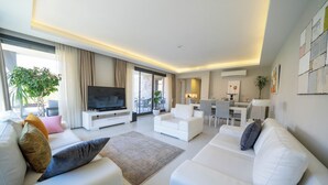 A modern living room with a flat-screen TV, white sofas, and a dining area with a table and chairs.