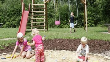 Kinder spielen in einer Sandspielanlage mit einer Holzschaukel und einer roten Rutsche.