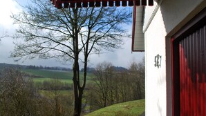 A building with a red garage door, a white wall with a number plaque, and a view of a tree-lined landscape with fields. - Holiday home in Reimboldshausen with balcony (Kirchheim/Seepark)