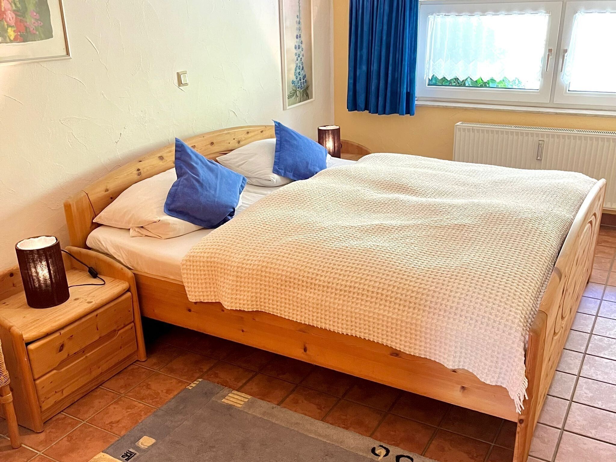 Una cama de madera con una colcha blanca, almohadas azules, una lámpara de noche y una mesita de noche.