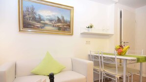 Een woonkamer met een witte bank, een groen kussen, een fles Belvilla wijn en twee glaasjes op een bijzettafeltje. Aan de muur hangt een schilderij en er is een zithoek met een tafel en stoelen.