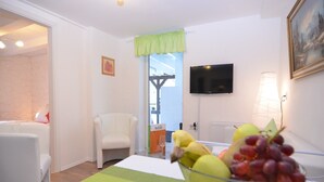 Een moderne woonkamer met een groen tapijt, een witte fauteuil, een glazen tafel met fruit, een flatscreen televisie en een schilderij aan de muur.