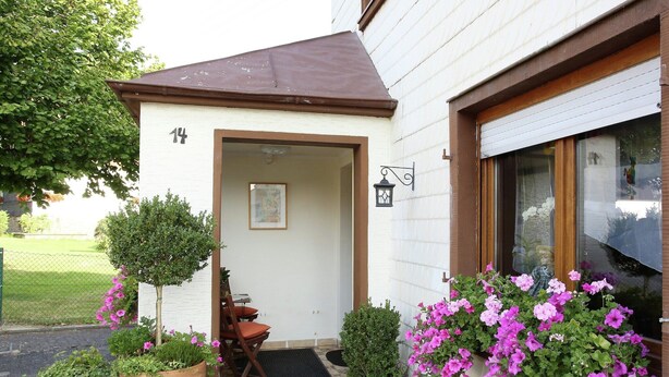 A white building with a brown roof, a door, and a window with pink flowers. There are potted plants and a small table with chairs outside.