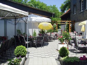 An outdoor café with tables and chairs, a yellow umbrella, and a white tent with text.