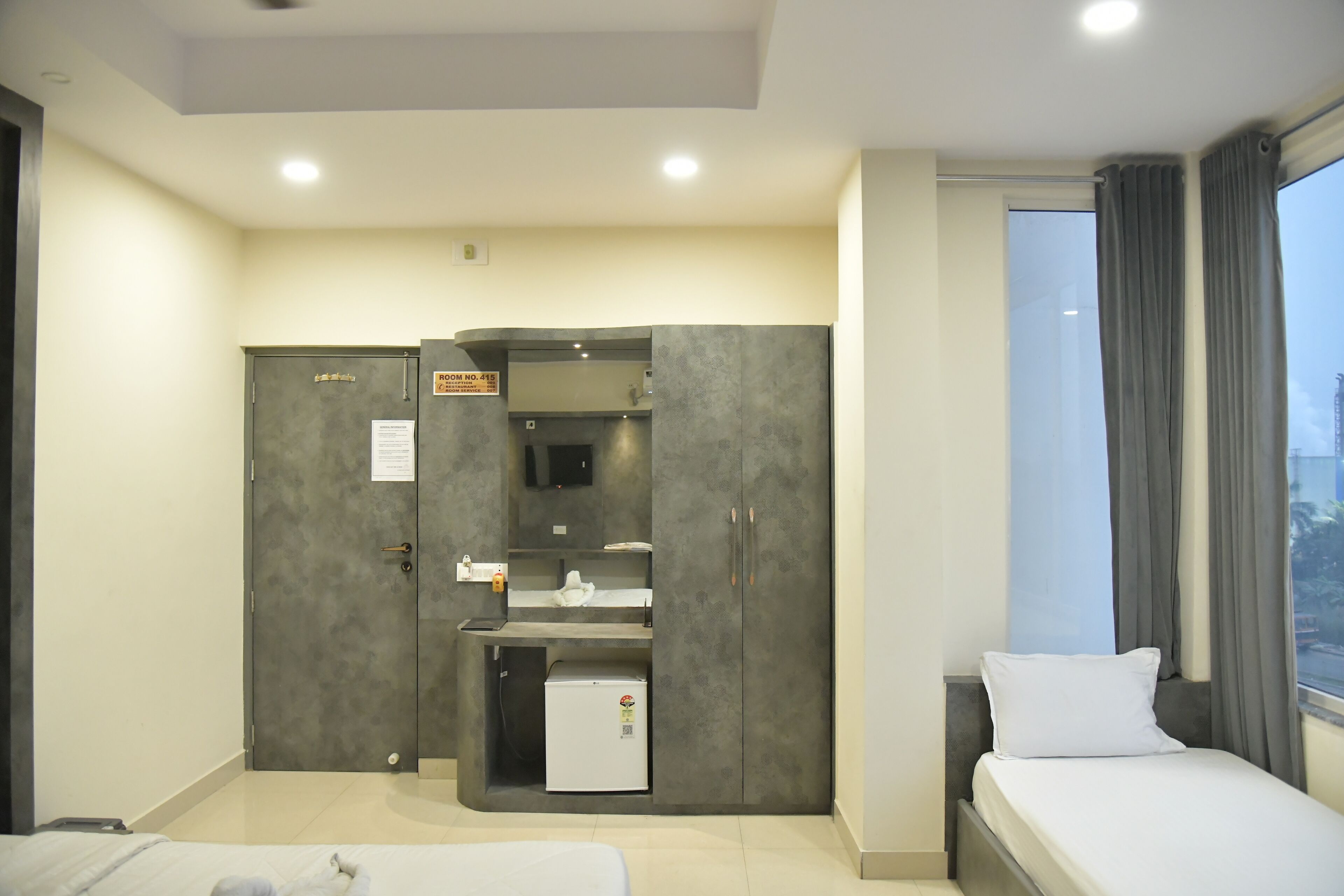 A hotel room with a bed, a desk with a refrigerator, a wardrobe, and a window with curtains.