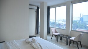 Een moderne hotelkamer met een groot bed, twee stoelen, een kleine tafel en uitzicht op de stad door grote ramen.