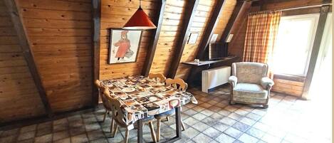 A cozy room with a wooden ceiling, a table with a patterned tablecloth, a chair, a hanging lamp, and a painting on the wall.