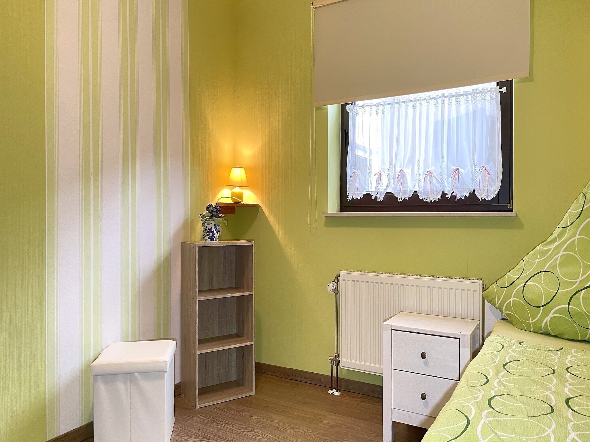 A bedroom with a bed, a bedside table, a nightstand, a lamp, a window with curtains, and a radiator.