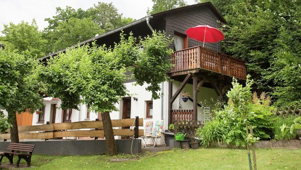 A two-story house with a balcony, surrounded by greenery and a wooden fence.