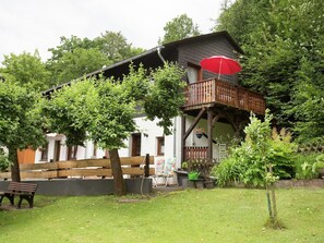 Uma casa de dois andares com varanda, rodeada por vegetação e um cercado de madeira.
