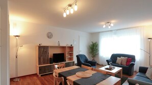Ein Wohnzimmer mit einem Holzesstisch, zwei Stühlen, einer Couch, einem Fernseher und einem Bücherregal.
