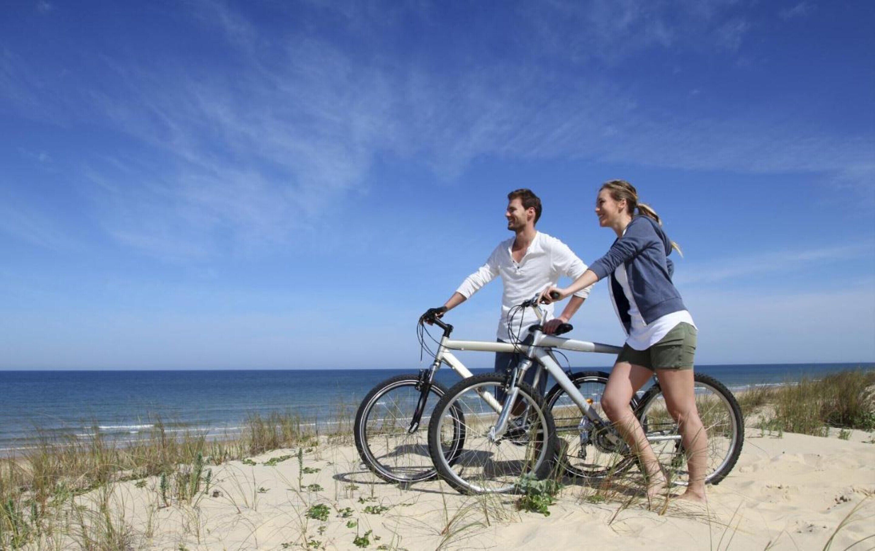 Deux personnes sur une plage, avec des vélos, des dunes de sable et l’océan en arrière-plan.