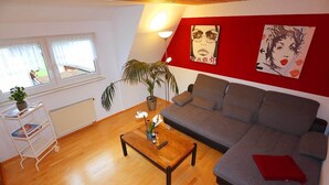 Ett vardagsrum med en grå soffa, ett träbord med blommor, en vit hylla med böcker, en krukväxt och en röd vägg med konstverk.