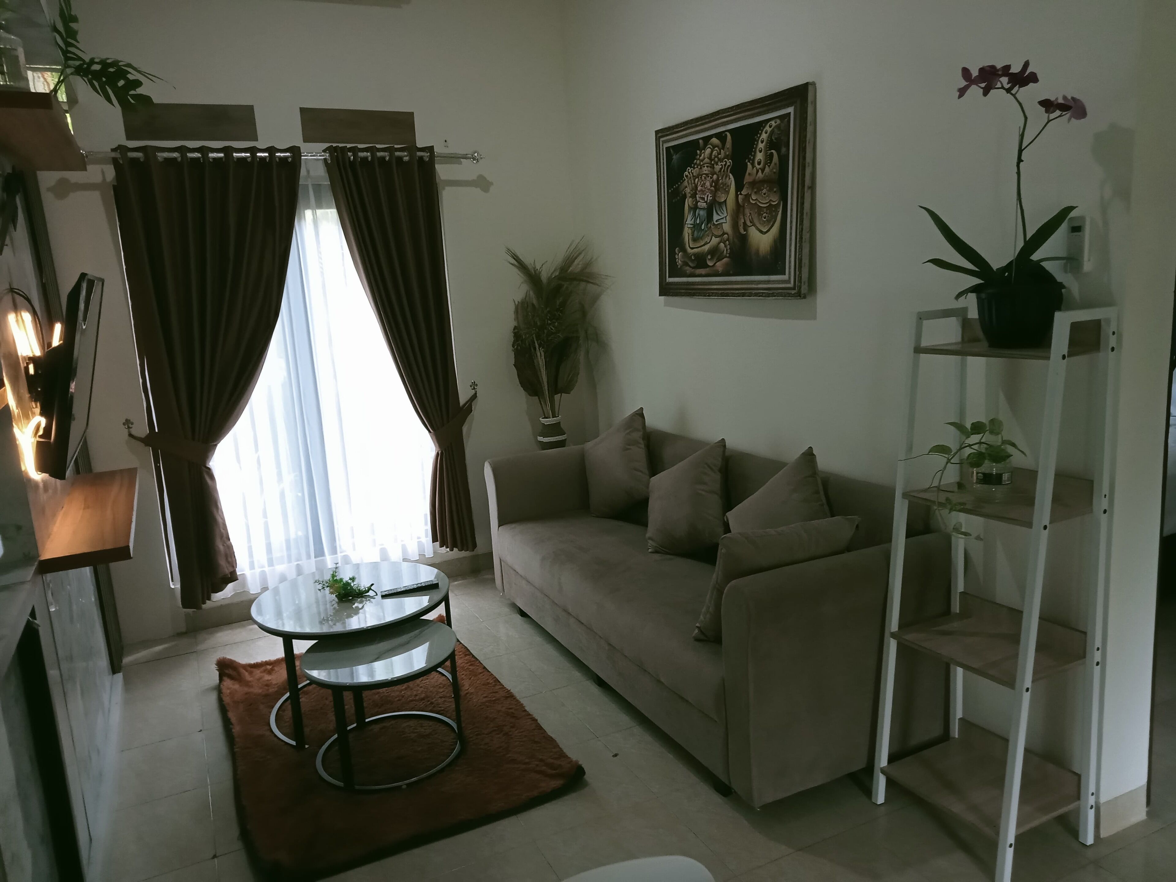 A living room with a sofa, a coffee table, a plant, a painting, and a shelf with more plants.