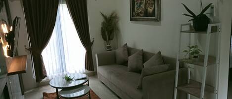 A living room with a sofa, a coffee table, a plant, a painting, and a shelf with more plants.