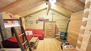 Ein gemütliches Dachzimmer mit Holzbalkendecke, einem roten Sofa, einer Holzleiter, einem kleinen Tisch und einem Korb mit Gegenständen.