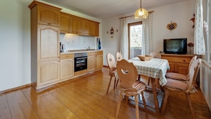 Een kleine keuken met houten kastjes, een inbouwoven en een eethoek met een tafel en stoelen.