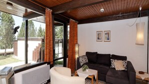 Una sala de estar moderna con un sofá oscuro, un sillón blanco y una mesita con una planta. Se observa una ventana grande con cortinas, un radiador y una lámpara de techo.