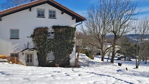 Una casa blanca con techo rojo, rodeada de nieve y árboles sin hojas.