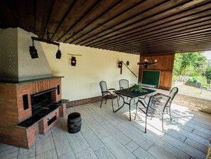 Patio esterno con caminetto in mattoni, sedie in metallo, un tavolo e uno schermo verde.