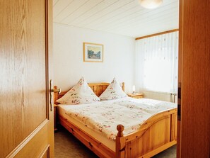 Un letto di legno con biancheria da letto floreale, un comodino con una lampada, un quadro appeso al muro e una finestra con tende leggere.